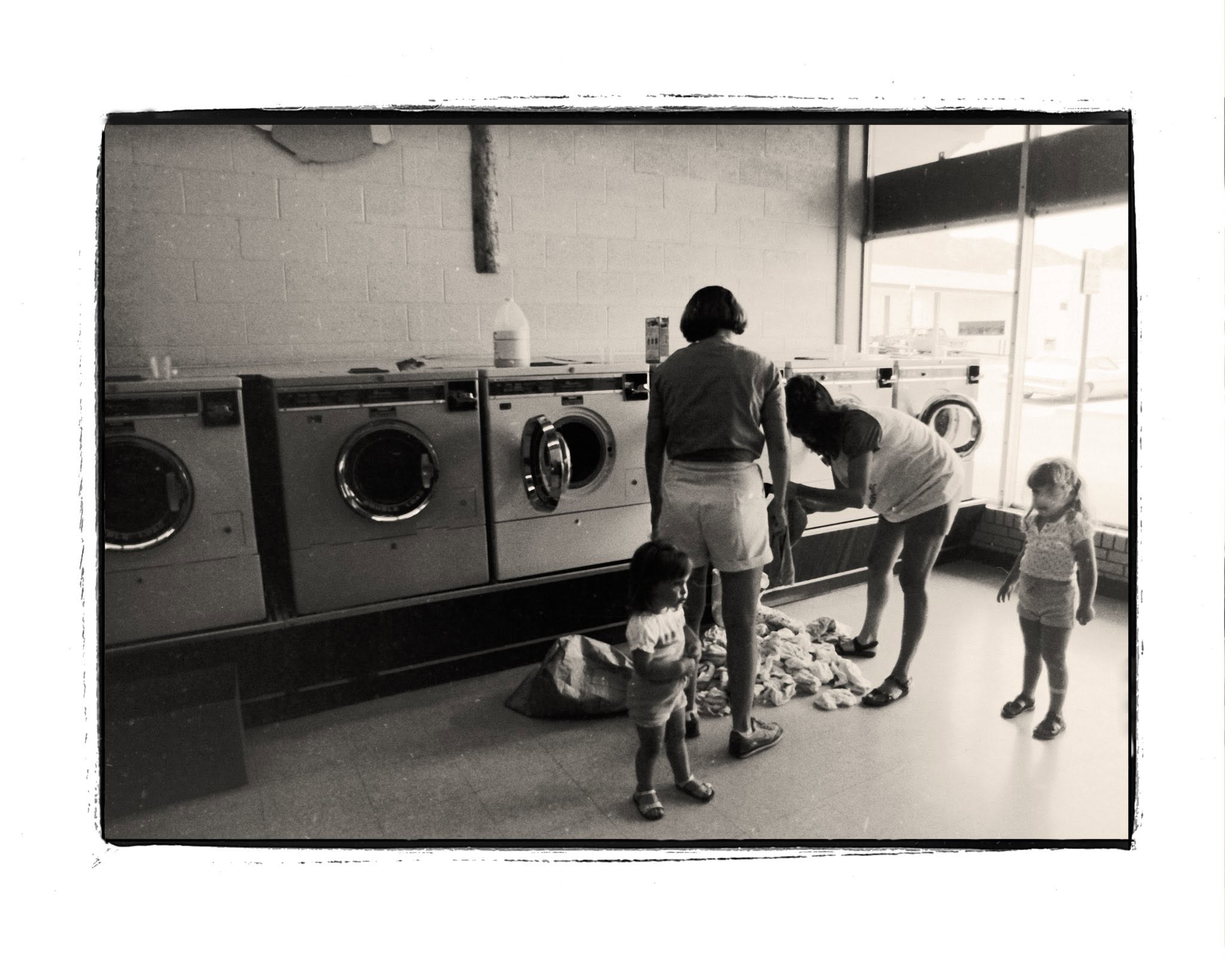 Three children and one adult stand in front of large washing machines at a laundromat, sorting clothes from a pile on the floor. Sunlight streams through large windows in the background.