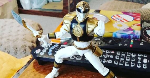An action figure of the White Power Ranger holding a sword is posed on a table cluttered with remote controls, tissues, and other household items.