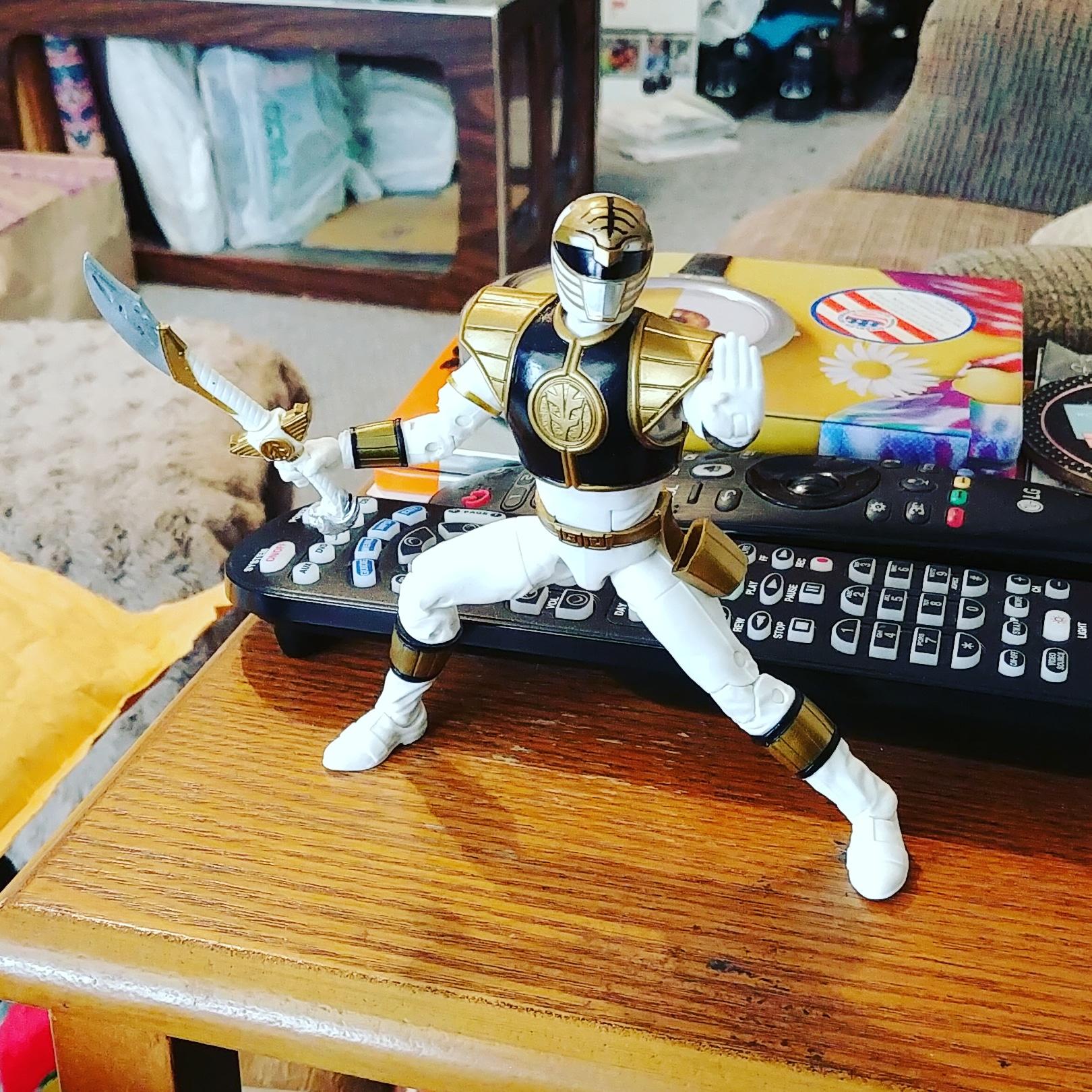 An action figure of the White Power Ranger is posed with a sword on a coffee table, standing on top of several TV remotes in a living room setting.