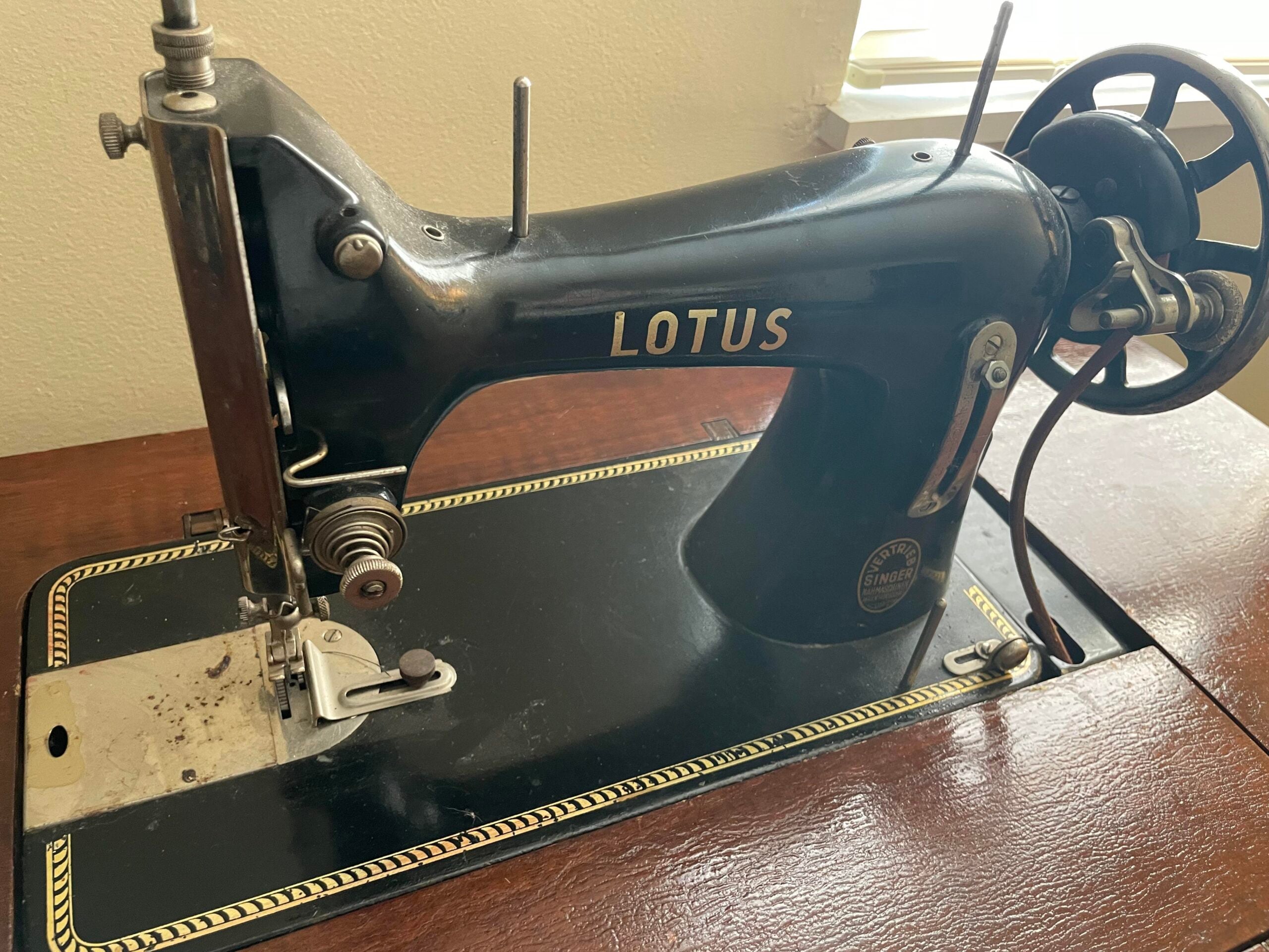 A vintage black Lotus sewing machine sits on a wooden table near a window, with gold decorative accents and visible metal parts, including a wheel and needle mechanism.