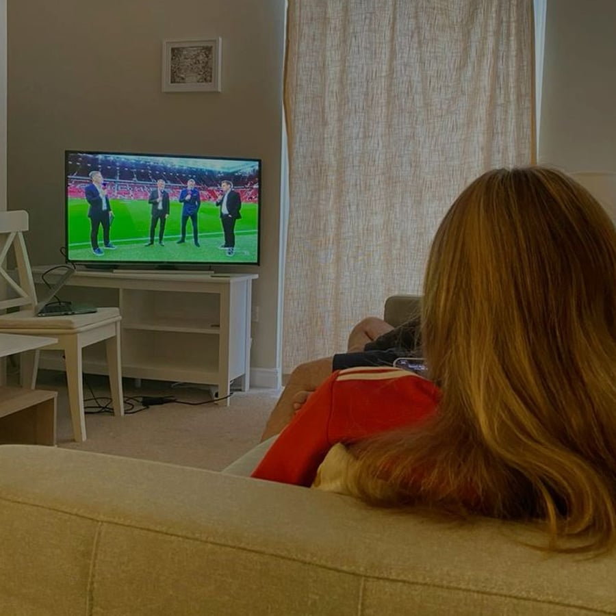 A person with long hair sits on a couch watching a soccer talk show on TV in a cozy living room with closed curtains and simple furniture.