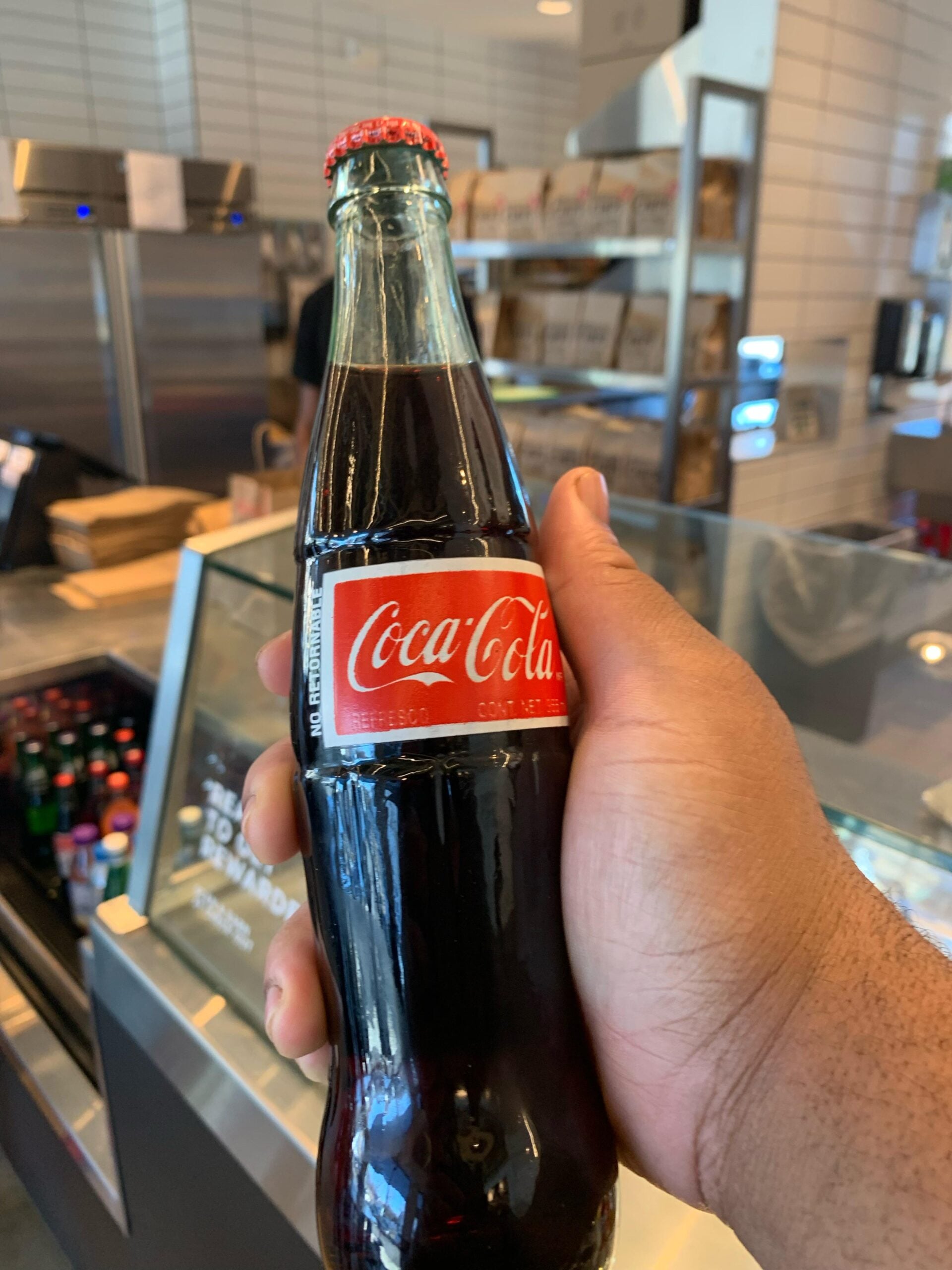 A hand holds a glass bottle of Coca-Cola inside a modern café, with shelves and a refrigerated display case in the background.