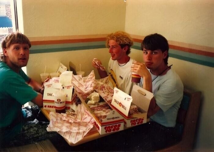 Three young men sit at a Taco Bell booth, eating and surrounded by numerous Taco Bell wrappers and drink cups on the table.