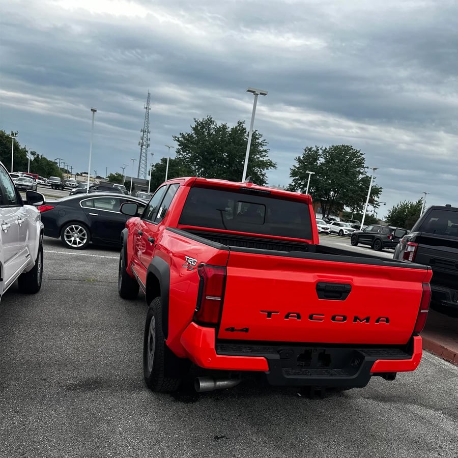 A red Toyota Tacoma pickup truck is parked in a lot among other cars on a cloudy day. Trees and light poles are visible in the background.