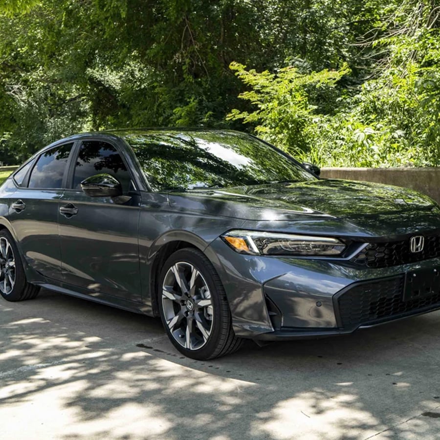 A dark gray Honda sedan parked on a shaded concrete driveway, surrounded by green trees and bushes on a sunny day.