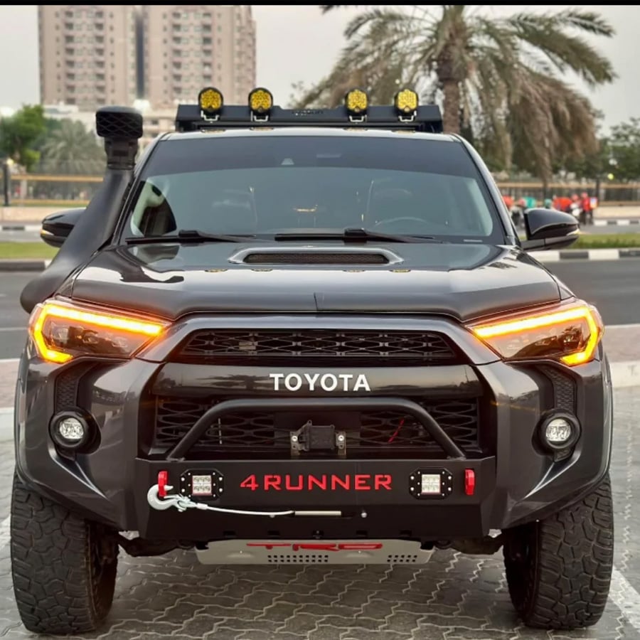 Front view of a black Toyota 4Runner SUV with off-road modifications, including a snorkel, roof lights, and a winch, parked on a street with palm trees and buildings in the background.