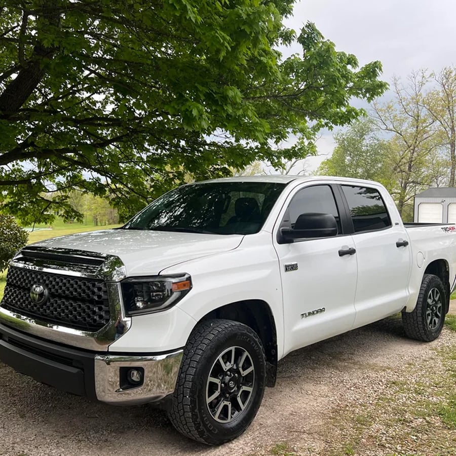A white Toyota Tundra pickup truck is parked on a gravel driveway under leafy green trees, with a small building and more trees visible in the background.