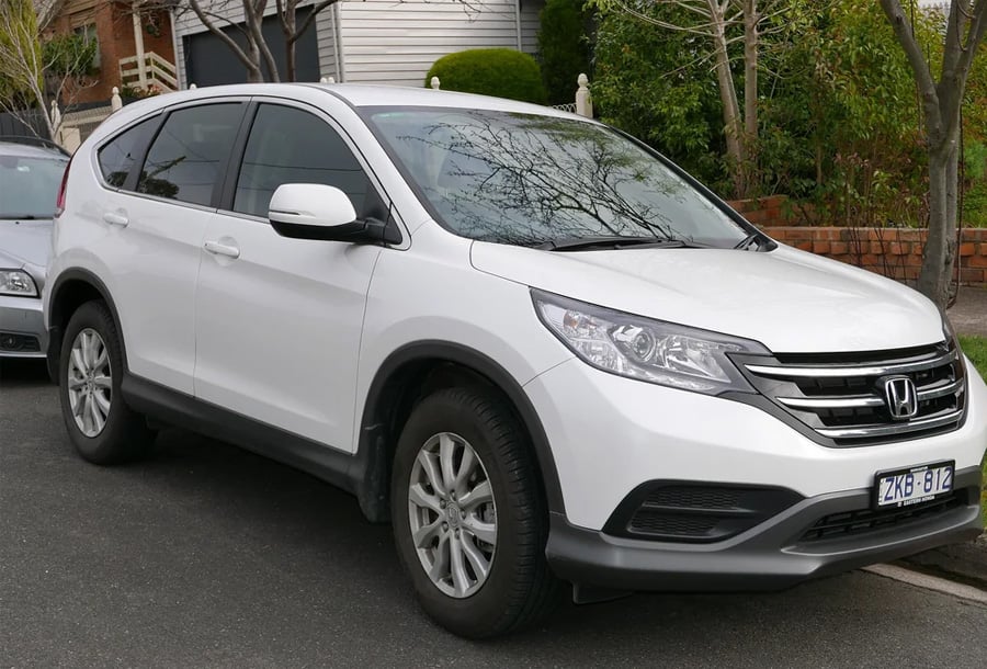 A white Honda CR-V SUV is parked on a residential street next to a curb, with trees and houses visible in the background.