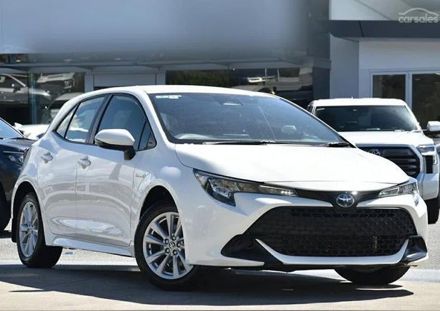 A white Toyota Corolla hatchback is parked outside a dealership on a sunny day, with other cars visible in the background.