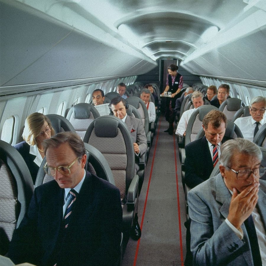 Passengers in business attire sit in rows inside an airplane cabin; a flight attendant stands in the aisle toward the back, serving drinks. Most people appear to be reading or working.