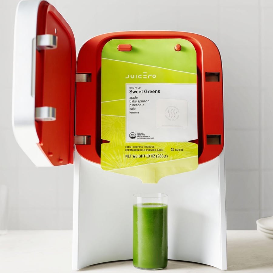 A Juicero machine squeezes a green juice packet labeled “Sweet Greens” into a glass, with the packet and glass clearly visible against a clean, white background.