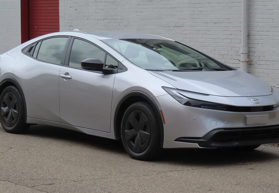 A silver Toyota Prius is parked on a paved surface near a white brick wall and a red garage door. The car has black wheel covers and a sleek, modern design.