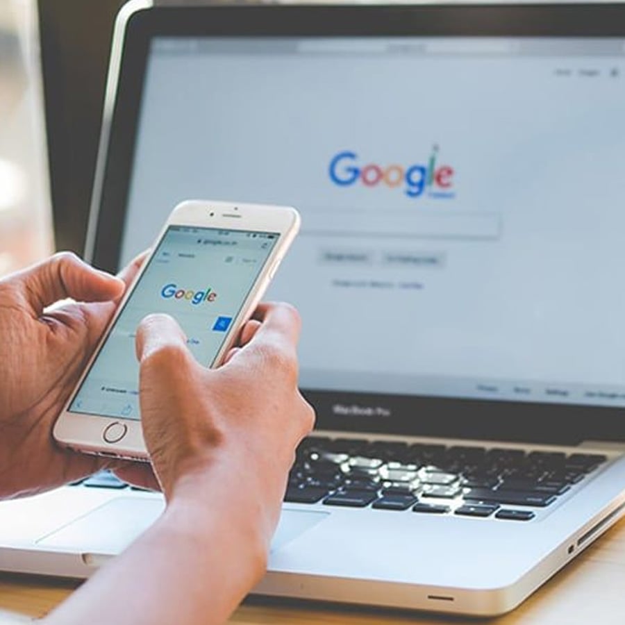 A person holds a smartphone with the Google search page open, while a laptop in the background also displays the Google homepage.
