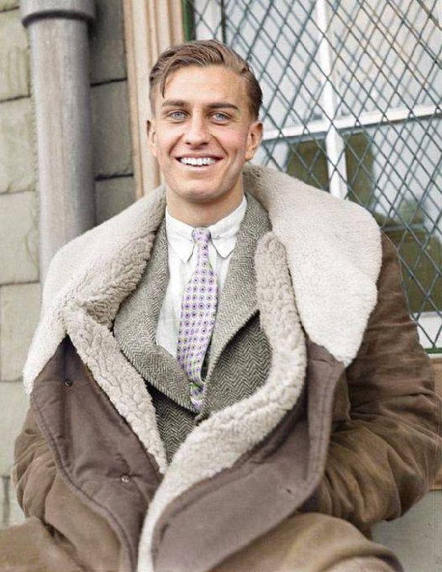 A young man in a vintage brown coat with a thick shearling collar sits and smiles, wearing a suit and patterned tie, in front of a leaded glass window.