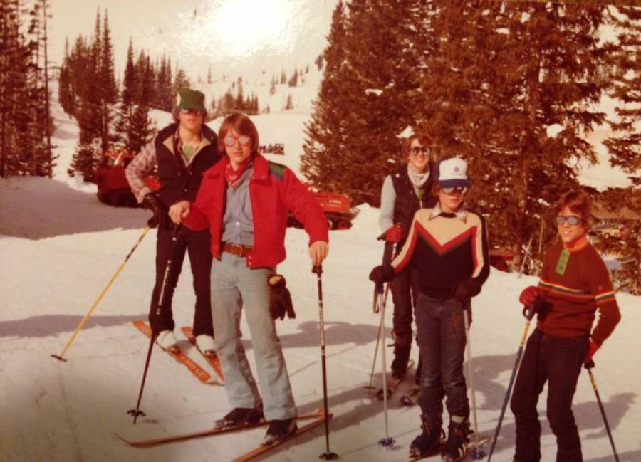 Five people stand on a snowy ski slope with trees and mountains in the background. They are dressed in retro ski outfits and holding ski poles, posing for the photo in bright daylight.