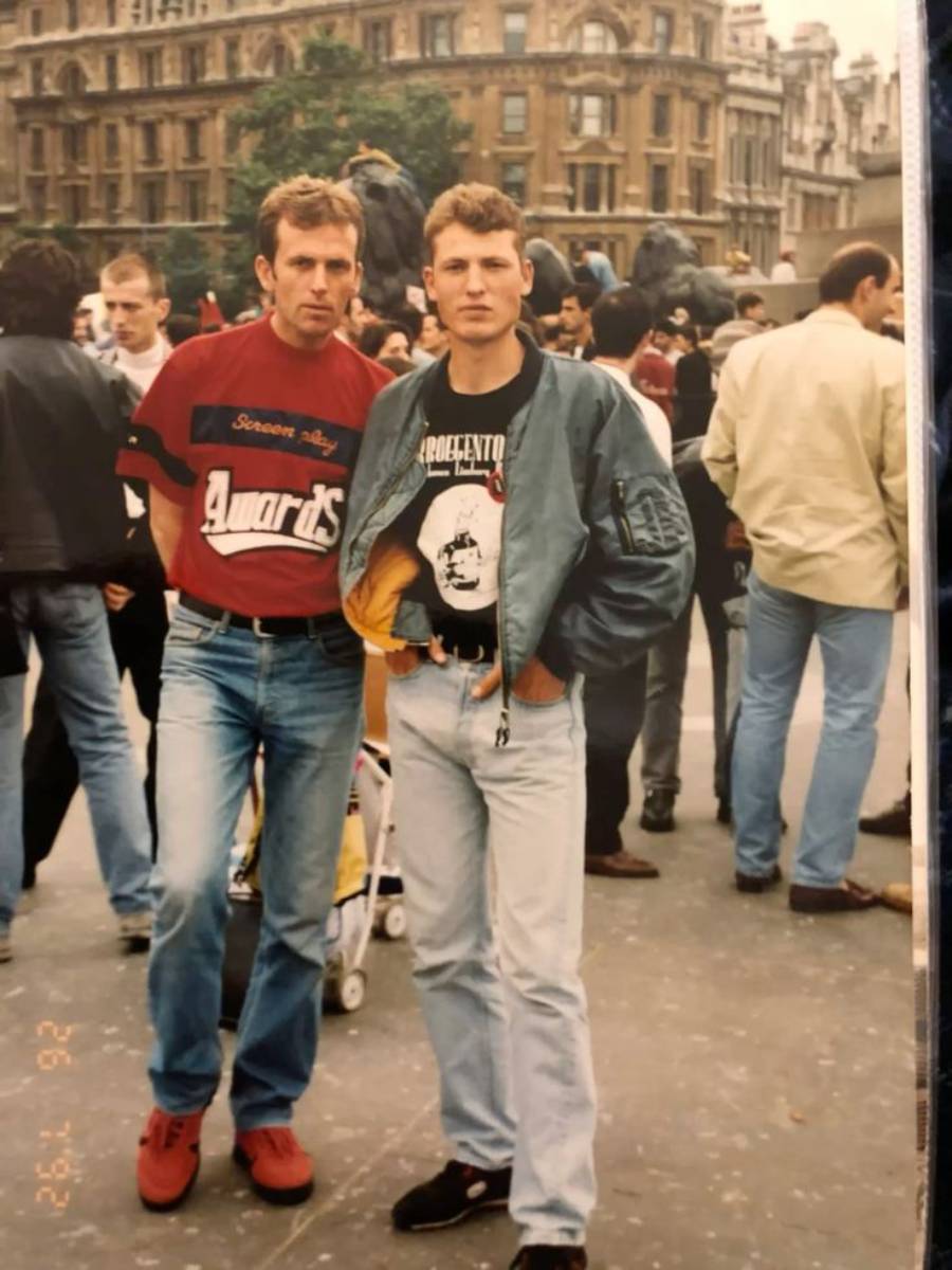 Two men pose in a busy city square, wearing casual clothes and jackets. People and historic buildings are visible in the background. One man has his hands in his pockets while the other stands with arms at his sides.