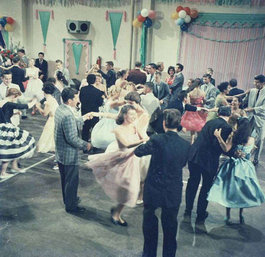 Teenagers in 1950s-style formal attire dance in a decorated gymnasium with balloons and streamers, creating a lively, colorful scene.