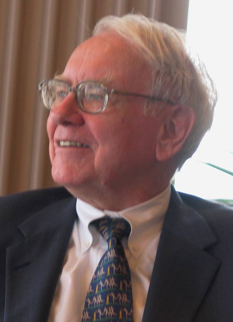 An older man with white hair and glasses, wearing a suit, white shirt, and patterned tie, is smiling and looking to the side in a warmly lit room with beige curtains in the background.