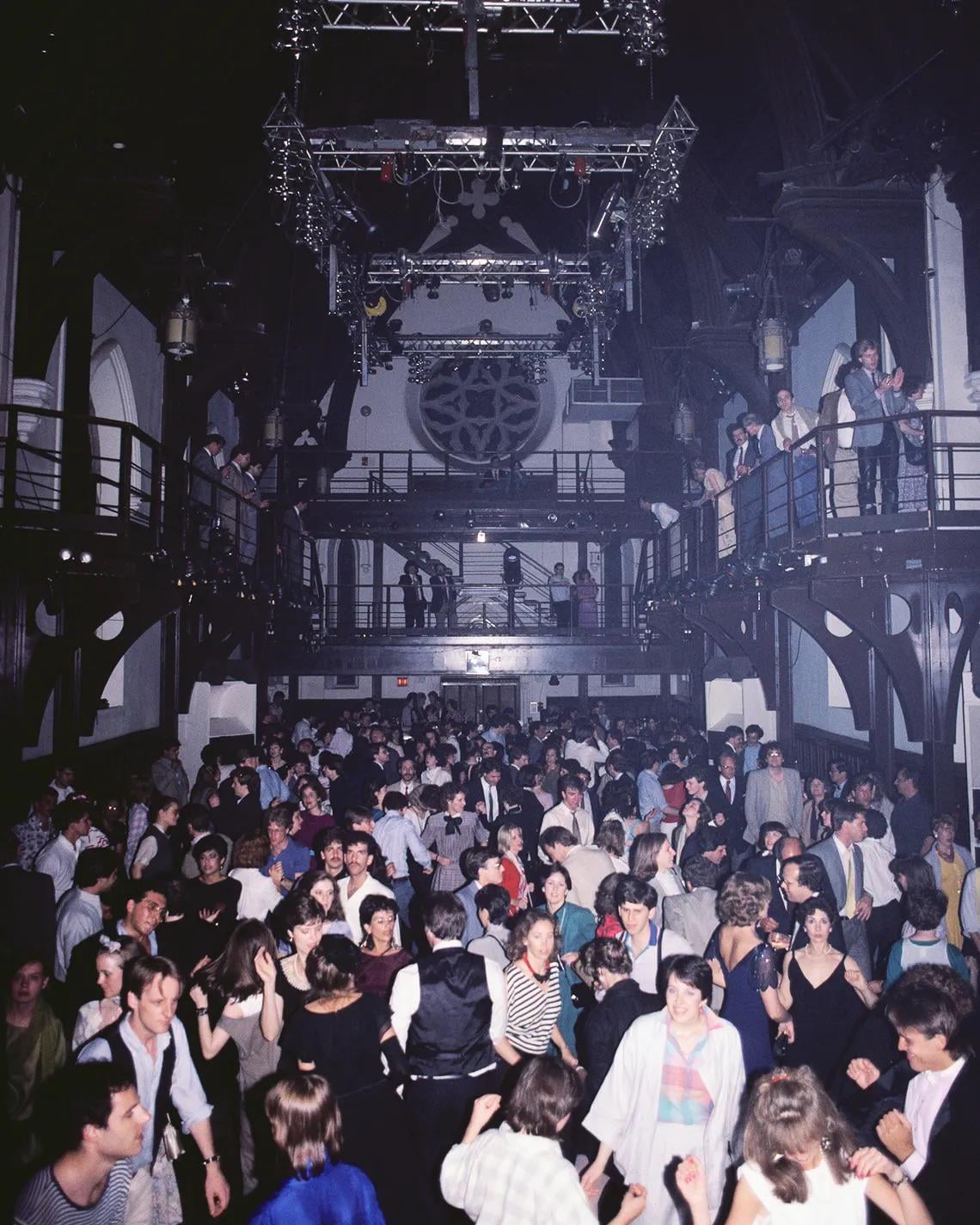 A large crowd of people dance and mingle in a packed, dimly lit nightclub with a high ceiling and balcony, while colorful lights shine overhead.