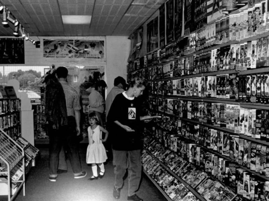 Black and white photo of people browsing VHS tapes in a video rental store. Shelves are lined with movie cases, and a child in a dress stands near some adults in the background.