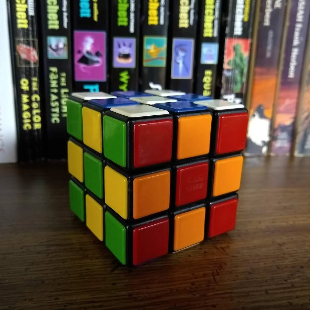 A partially solved Rubik’s Cube sits on a wooden surface in front of a row of colorful books. The cube’s green, yellow, orange, and red sides are visible.