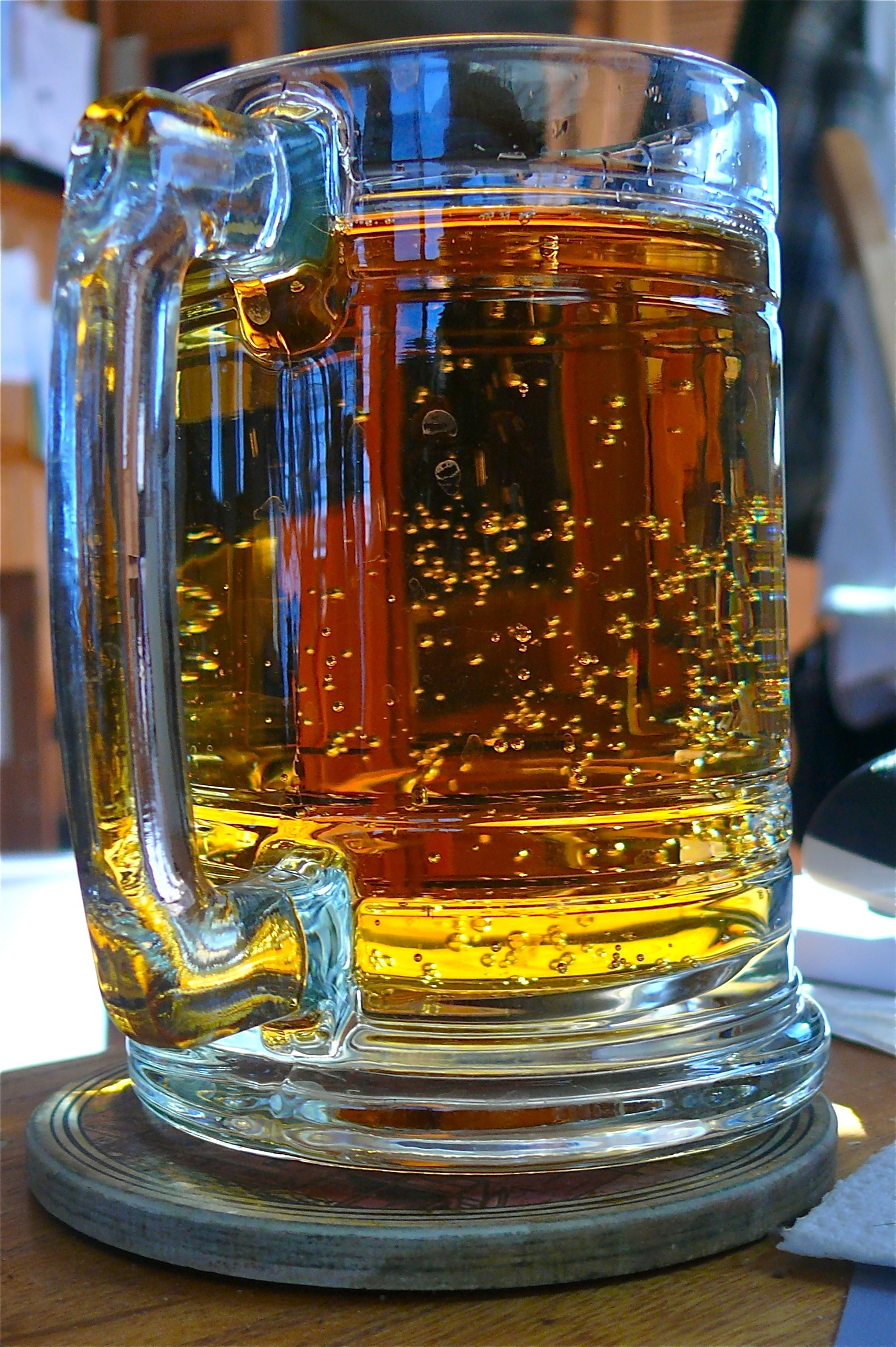 A close-up of a glass mug filled with amber-colored beer, showing bubbles rising inside, resting on a wooden coaster in natural sunlight.