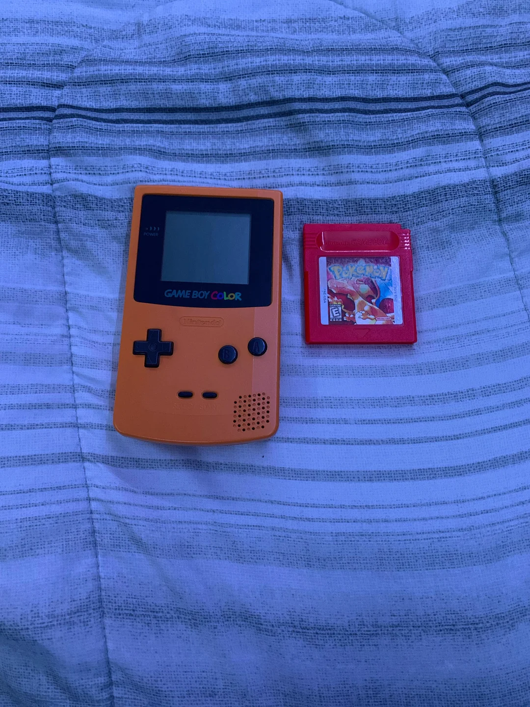 An orange Game Boy Color and a red Pokémon game cartridge are placed side by side on a striped fabric background.