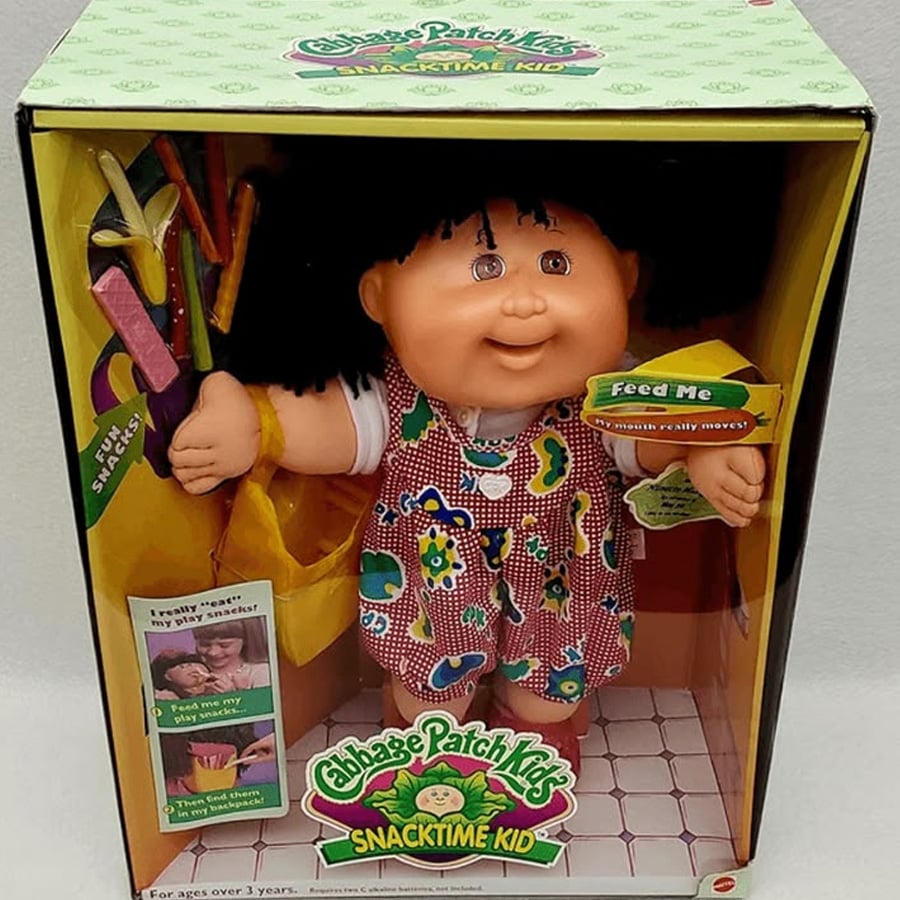 A Cabbage Patch Kids Snacktime Kid doll in a box, wearing a colorful dress with a yellow bag and snacks. The box has bright colors and text, with the doll’s arm raised and curly black hair.
