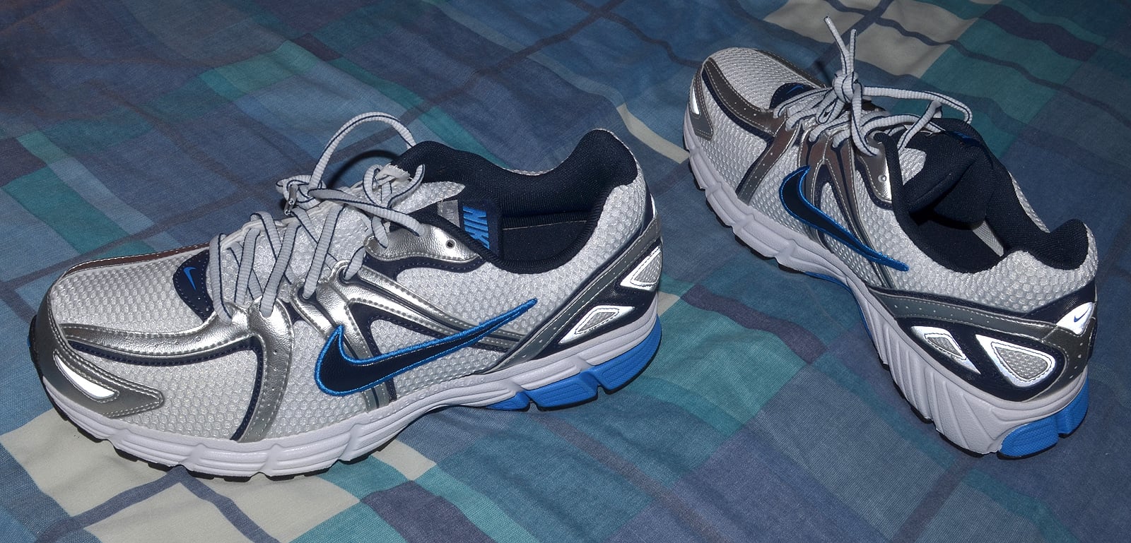 A pair of gray and silver Nike running shoes with blue accents are placed on a blue and green plaid bedspread.