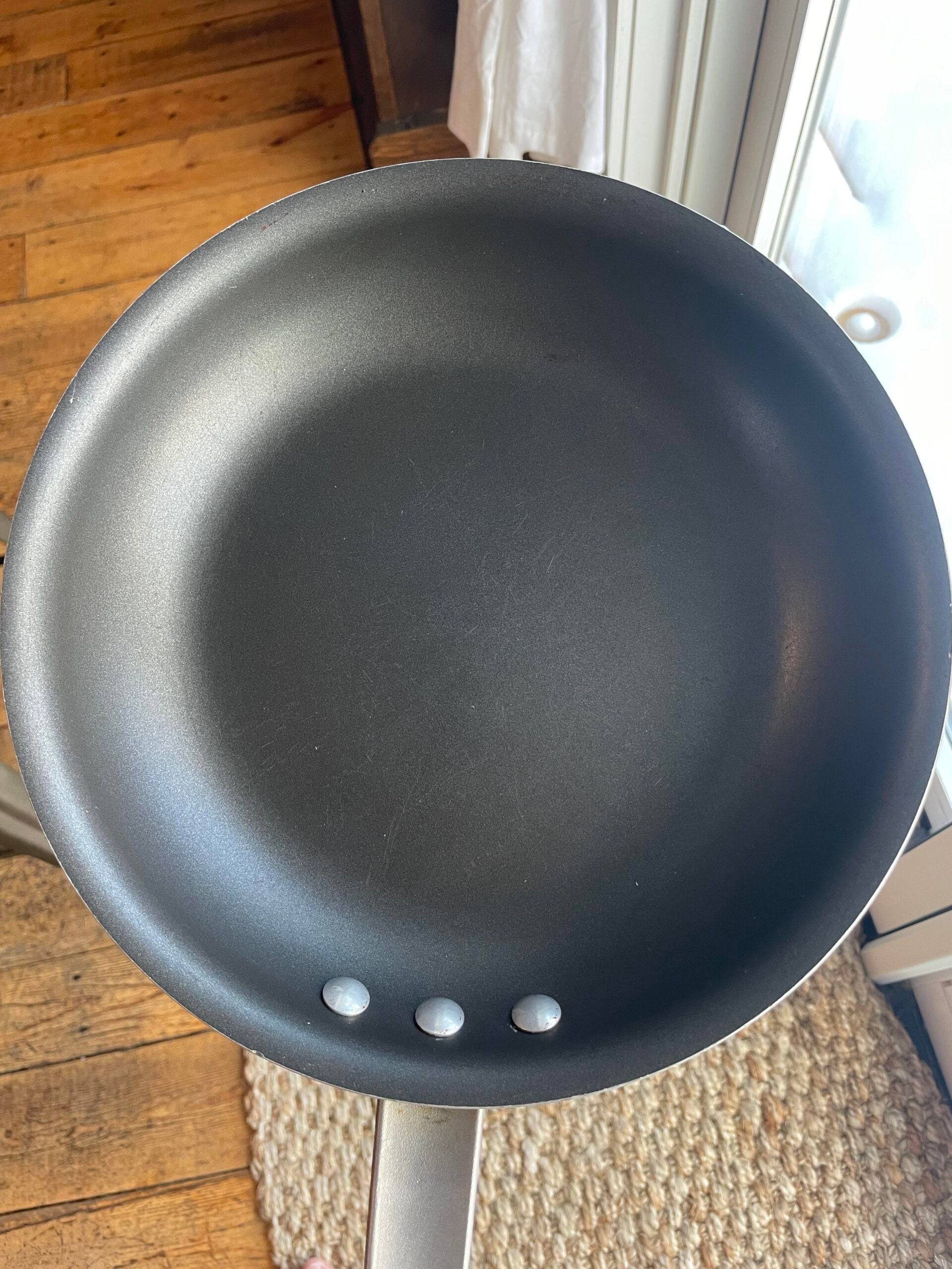 A black nonstick frying pan with three rivets is held above a wooden floor and a woven rug, near a glass door letting in natural light.
