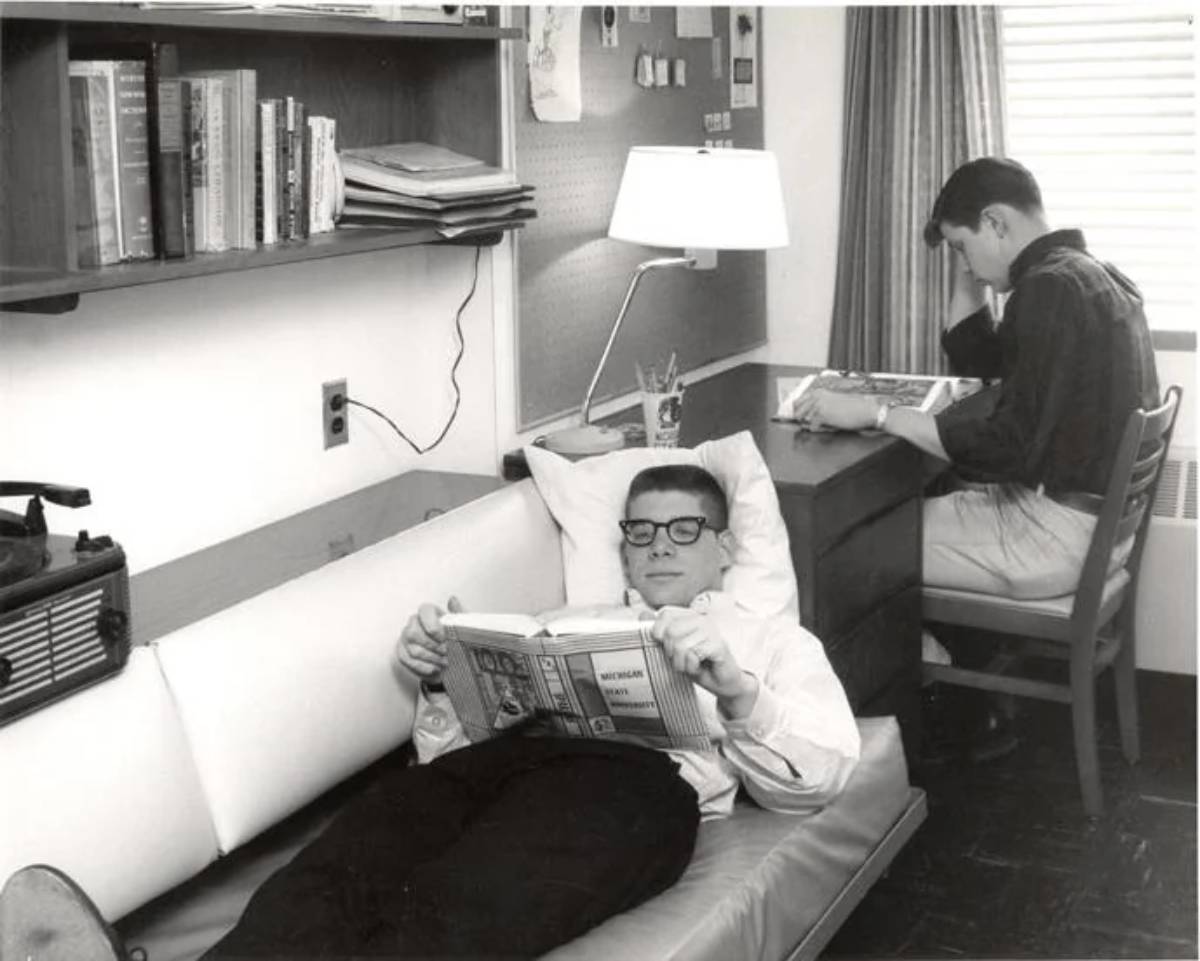 Two young men study in a dorm room; one reclines on a couch reading a book, while the other sits at a desk writing. Books, a lamp, and a window with blinds are visible in the background.