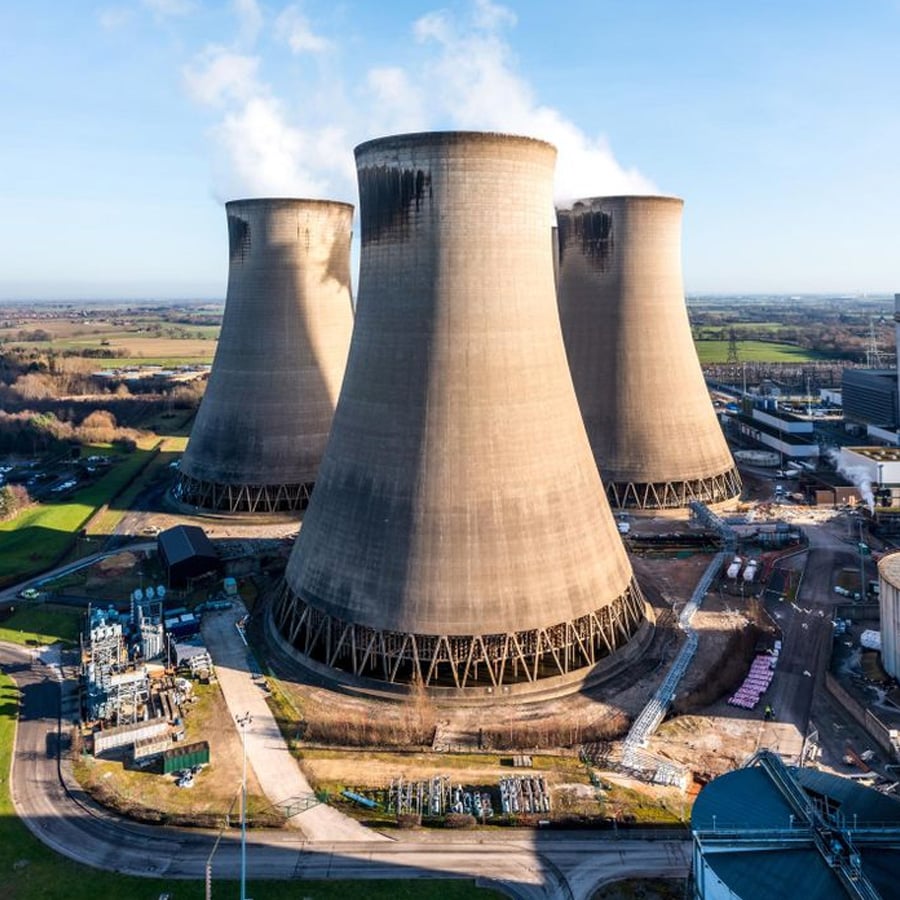 Aerial view of four large cooling towers at a power plant, with steam rising and surrounding industrial buildings, set in a rural landscape under a clear blue sky.