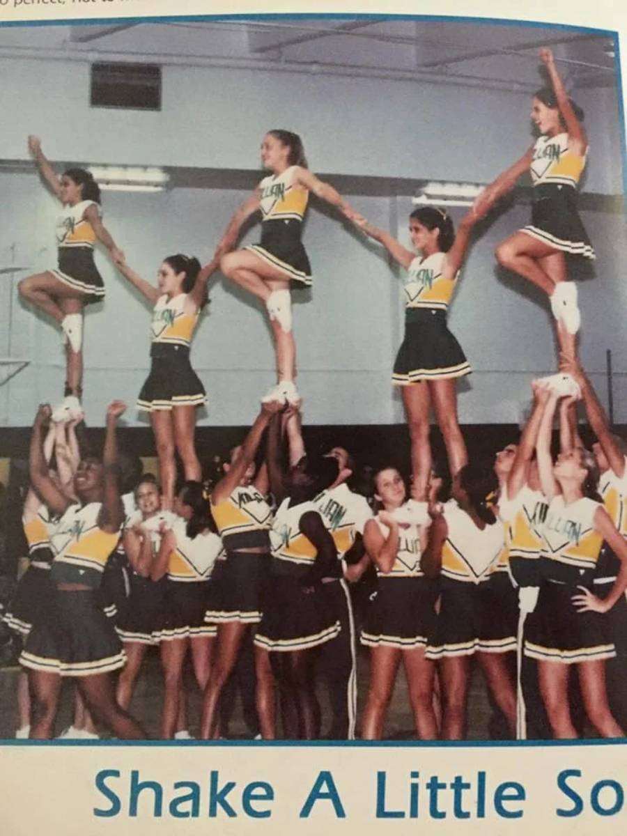 A cheerleading team in yellow, green, and white uniforms performs a stunt, lifting four members high with arms raised. The scene takes place in an indoor gymnasium.