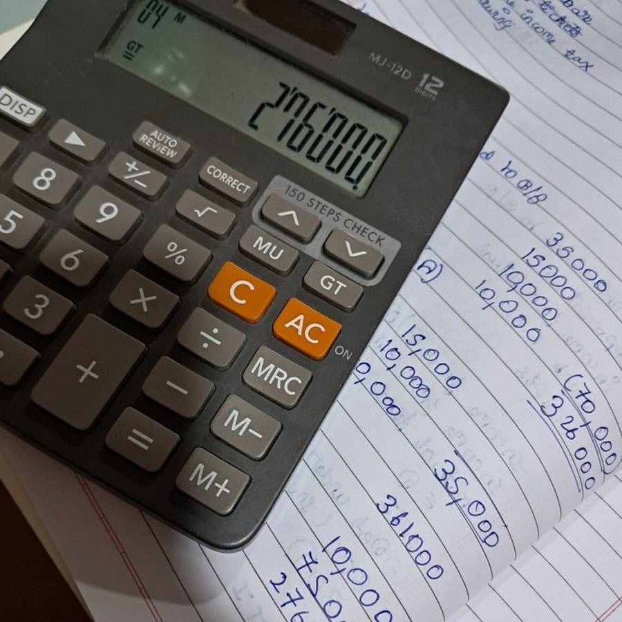A calculator displaying “276000” rests on an open notebook filled with handwritten numerical calculations in blue ink.