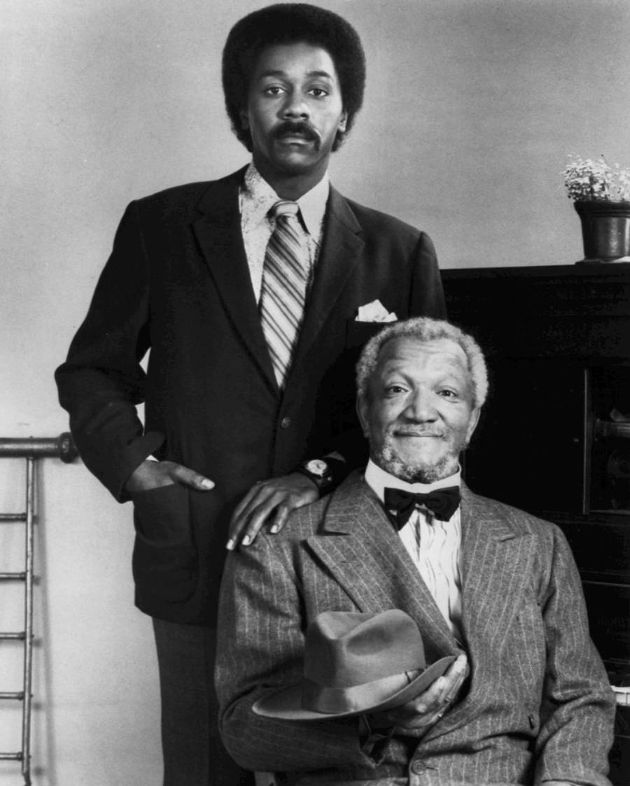 Two men in suits pose for a black-and-white portrait; one stands with his hand on a chair, the other sits holding a hat and smiling, with a piano and small plant in the background.