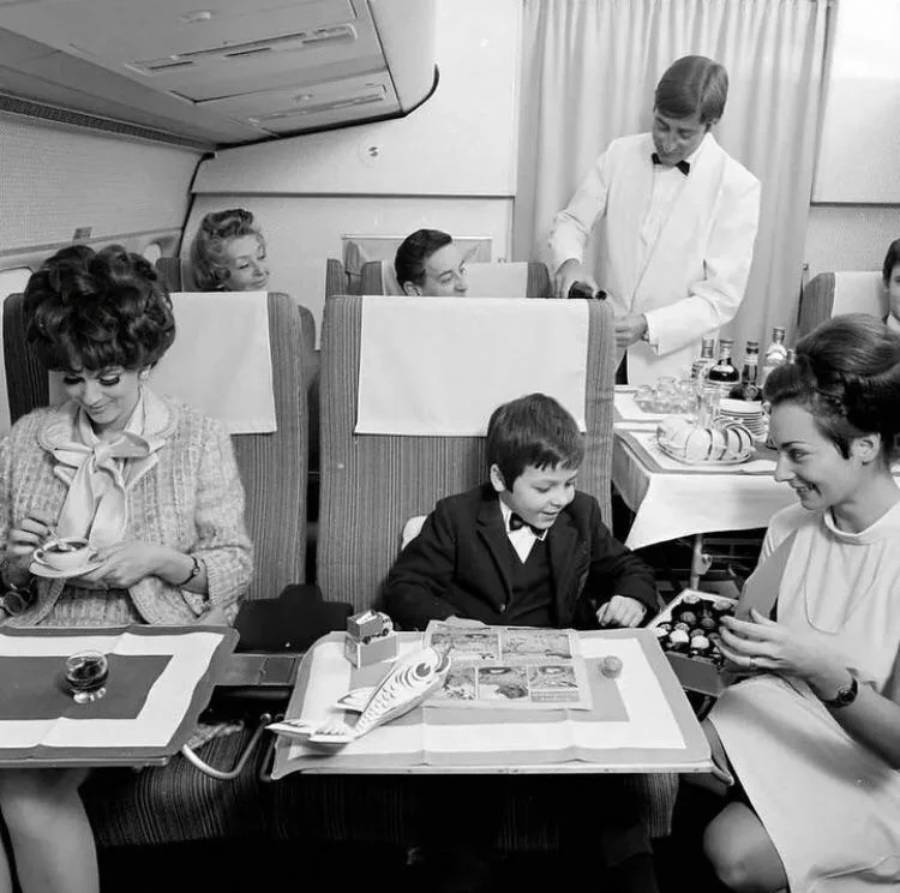 Black and white photo of passengers, including a child, enjoying a meal in airplane seats. A flight attendant in a white jacket serves drinks. The scene appears to be from the mid-20th century.