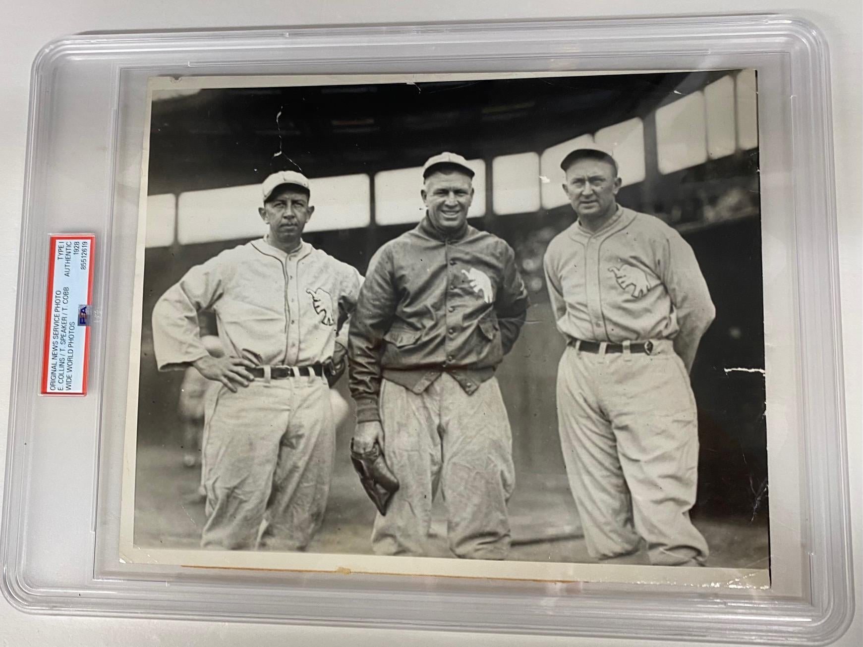 Three baseball players in vintage uniforms pose together on a field. The black-and-white photo is encased in a protective plastic holder. The background shows stadium seating and large windows.