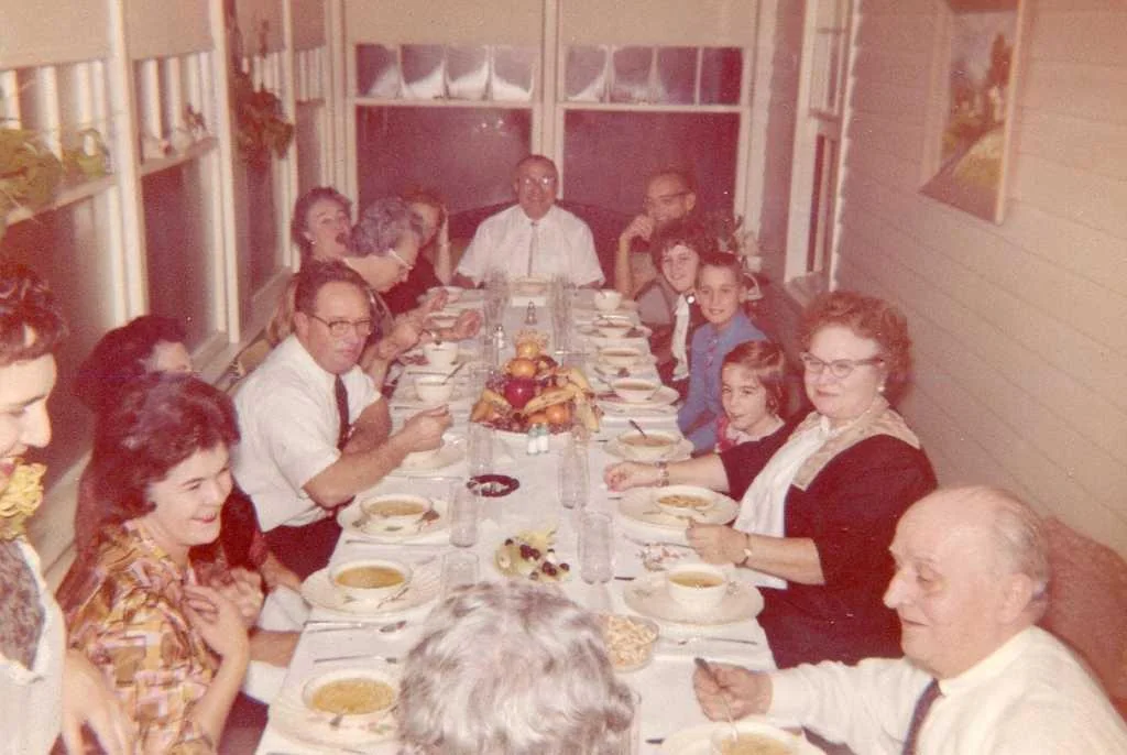 A large family sits around a long table set with soup bowls and bread, sharing a meal together in a cozy, sunlit room. Smiles and conversation suggest a warm gathering, possibly a holiday or special occasion.