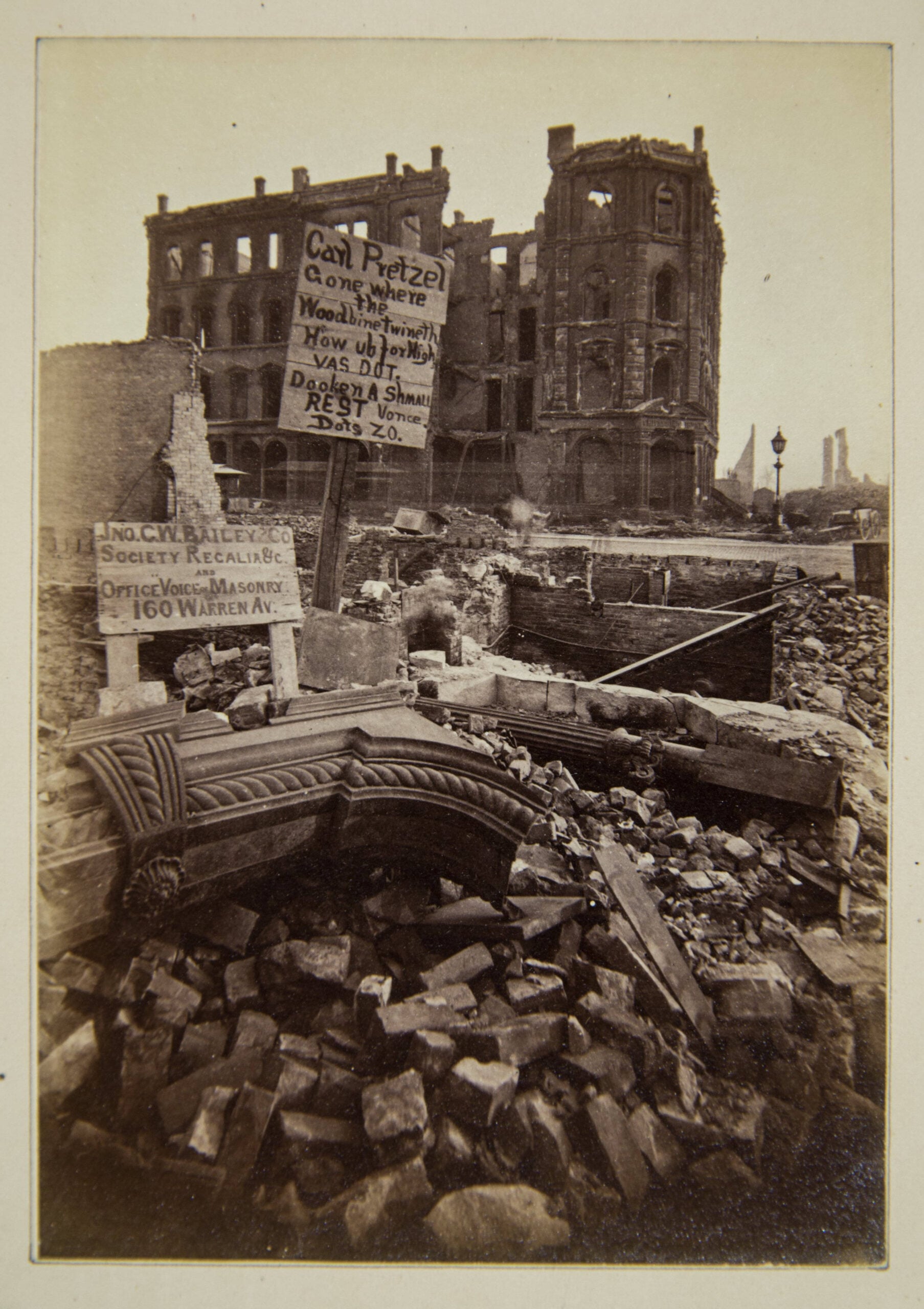 A ruined building stands amid rubble. Two signs offer pretzels and society relief support, with handwritten messages atop the debris. The scene is desolate and marked by the aftermath of destruction.