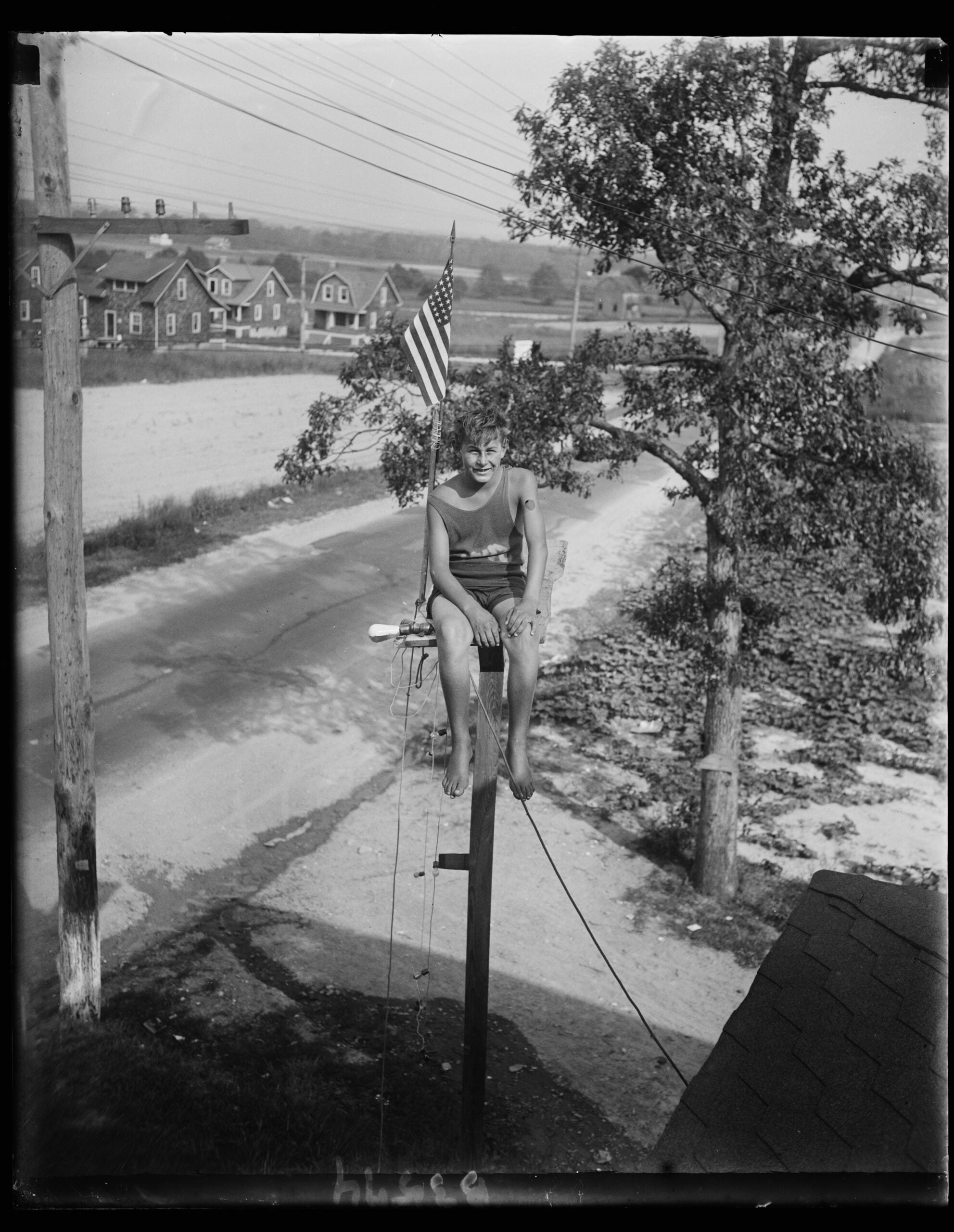 A person in a swimsuit sits atop a tall pole holding an American flag. There’s a road, trees, power lines, and houses in the background. The scene appears to be from an earlier era.