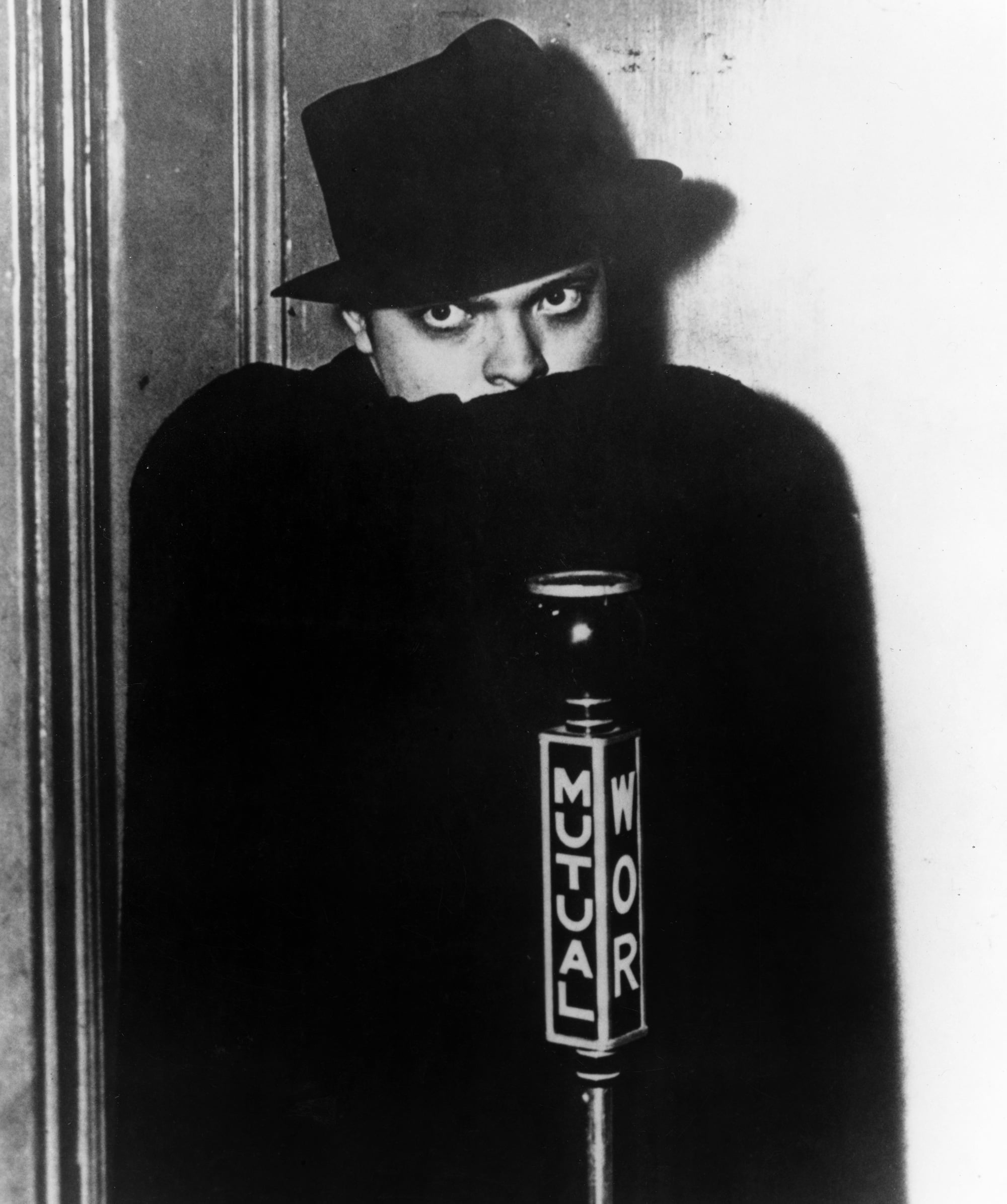 A person in a dark cloak and hat stands behind a vintage microphone labeled “Mutual WOR,” with only their eyes and nose visible, looking directly at the camera.