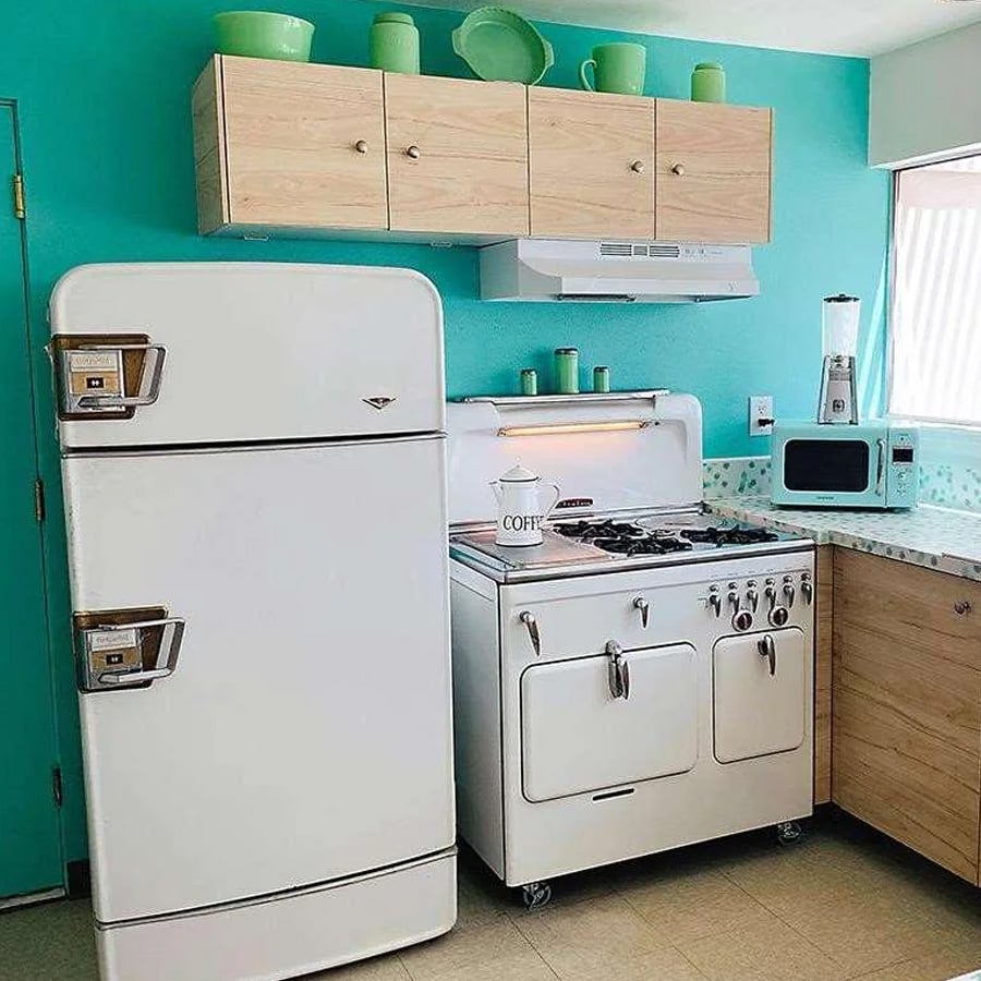A retro kitchen with a vintage white fridge and stove, turquoise walls, wooden cabinets, mint green dishes above, a coffee pot on the stove, and a blender on the counter by the window.