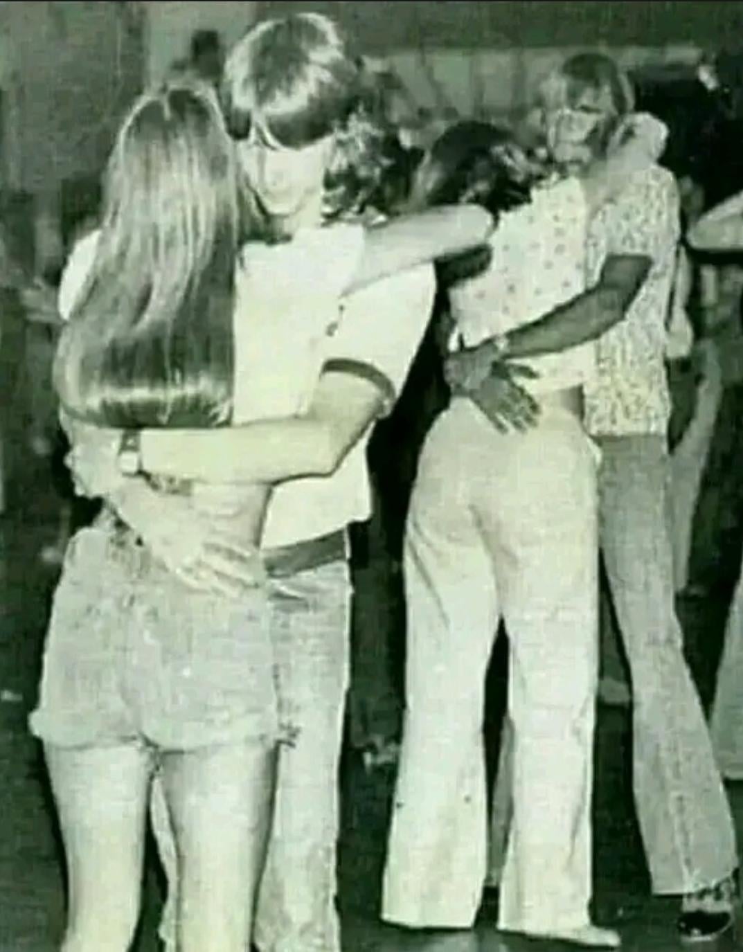 Two young couples slow dance closely in a dimly lit room. Both couples have their arms wrapped around each other, and the scene appears to be from the 1970s, based on their hairstyles and clothing.