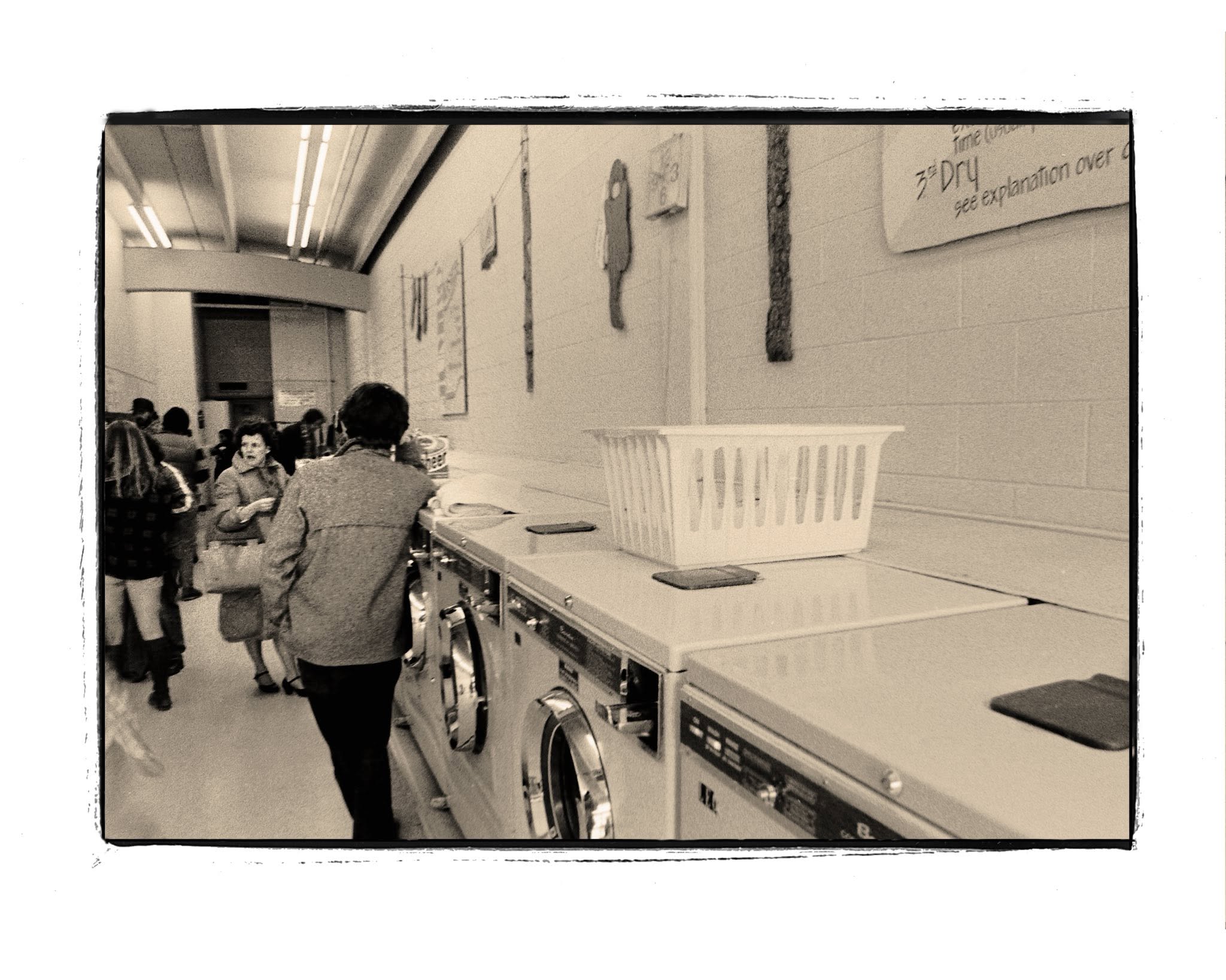 Sepia-toned photo of people in a laundromat. A plastic laundry basket sits on top of washing machines. Art and signs hang on the wall. Several people walk down the aisle.