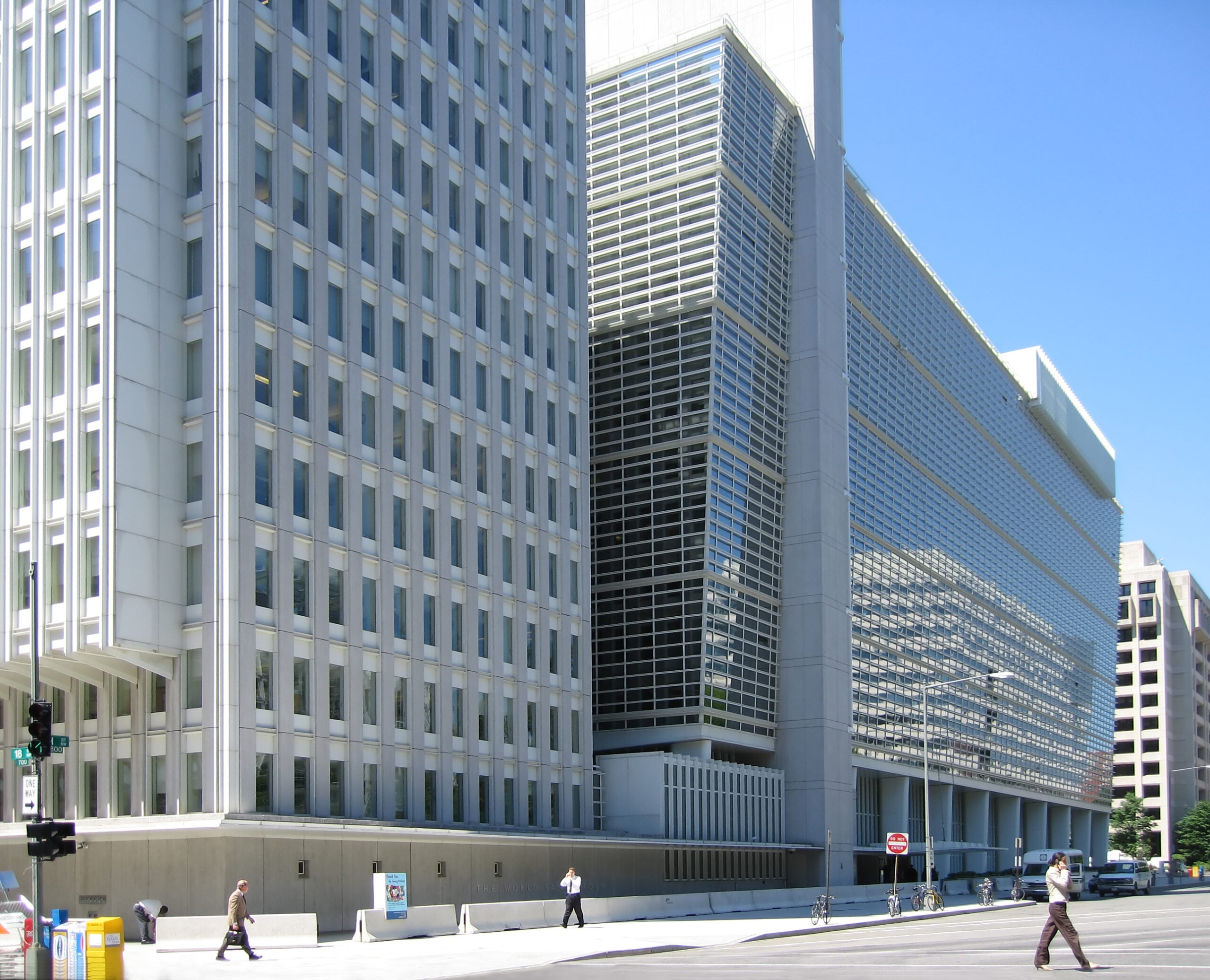 Modern office buildings with glass and steel facades, seen on a sunny day. Several people are walking on the sidewalk in front, and a few cars are parked along the street in the foreground.