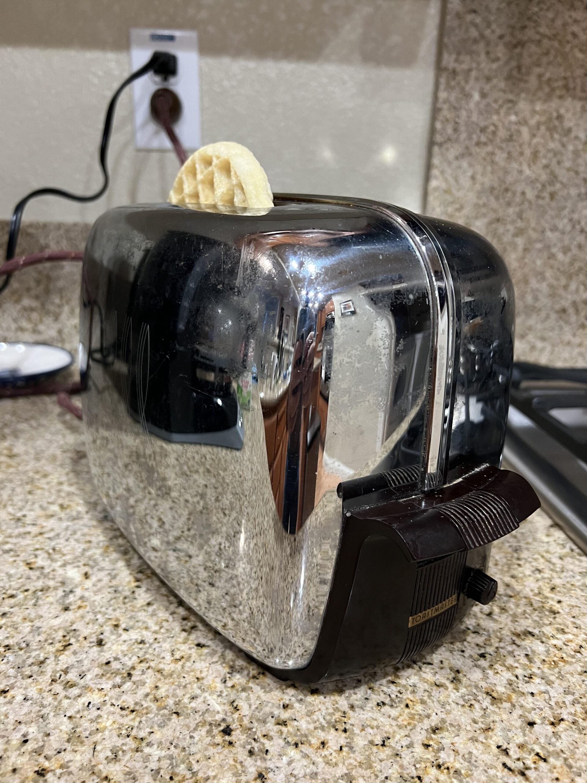 A small waffle is sticking out of a shiny chrome toaster on a kitchen counter. The toaster’s reflection shows part of the kitchen.