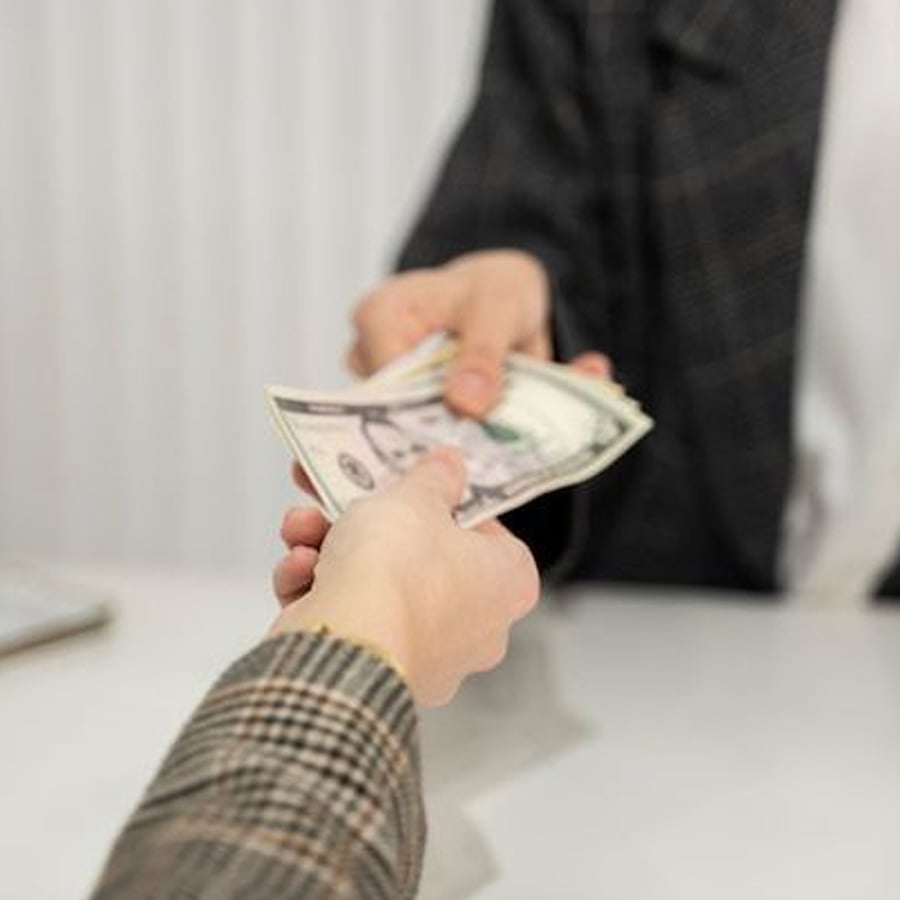 Two people, both wearing plaid sleeves, exchange a stack of US dollar bills across a white table, with one hand giving and the other receiving the money.