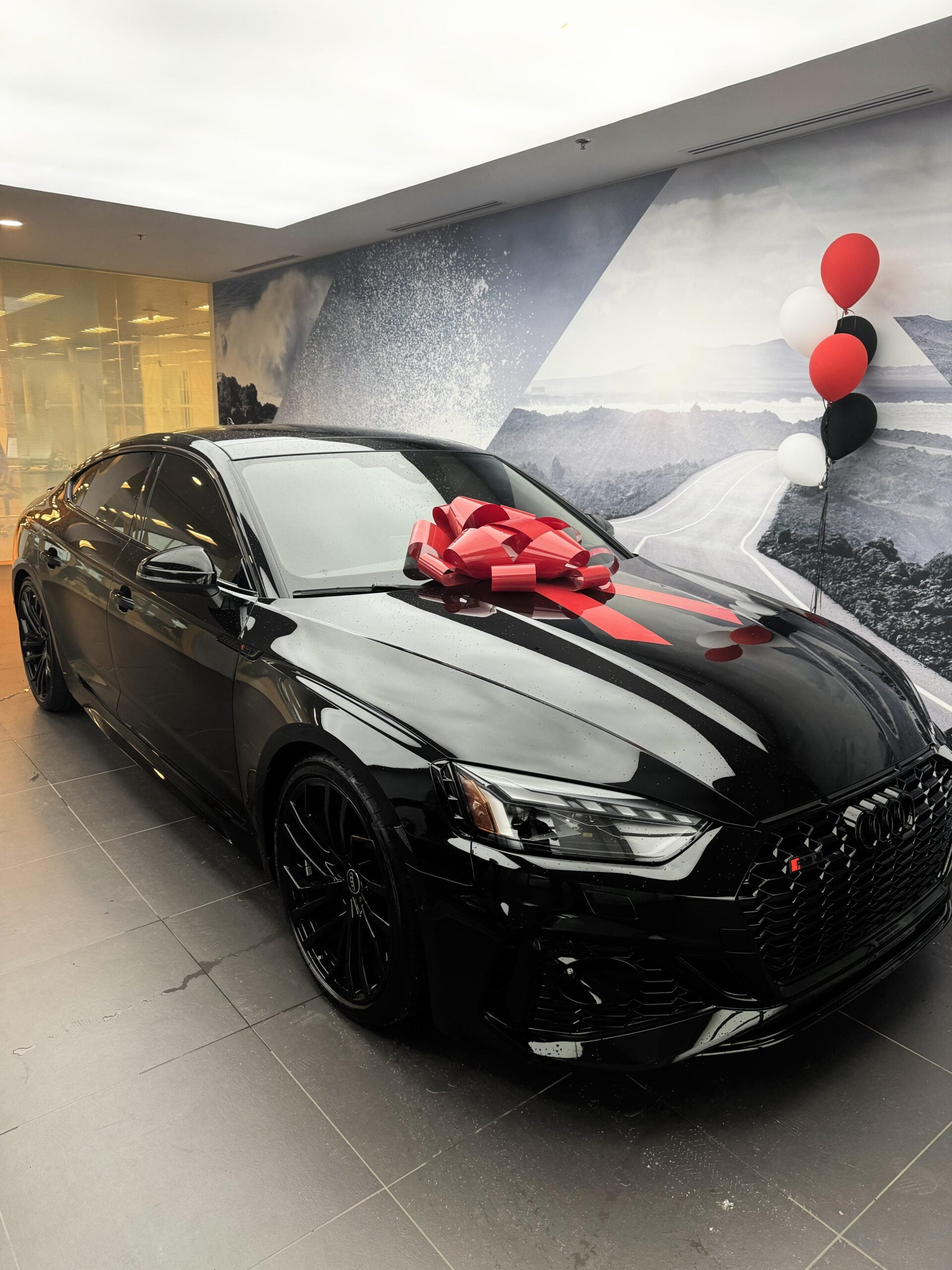 A shiny black Audi sedan with a large red bow on the hood and red, white, and black balloons attached, displayed indoors near a scenic mural background.
