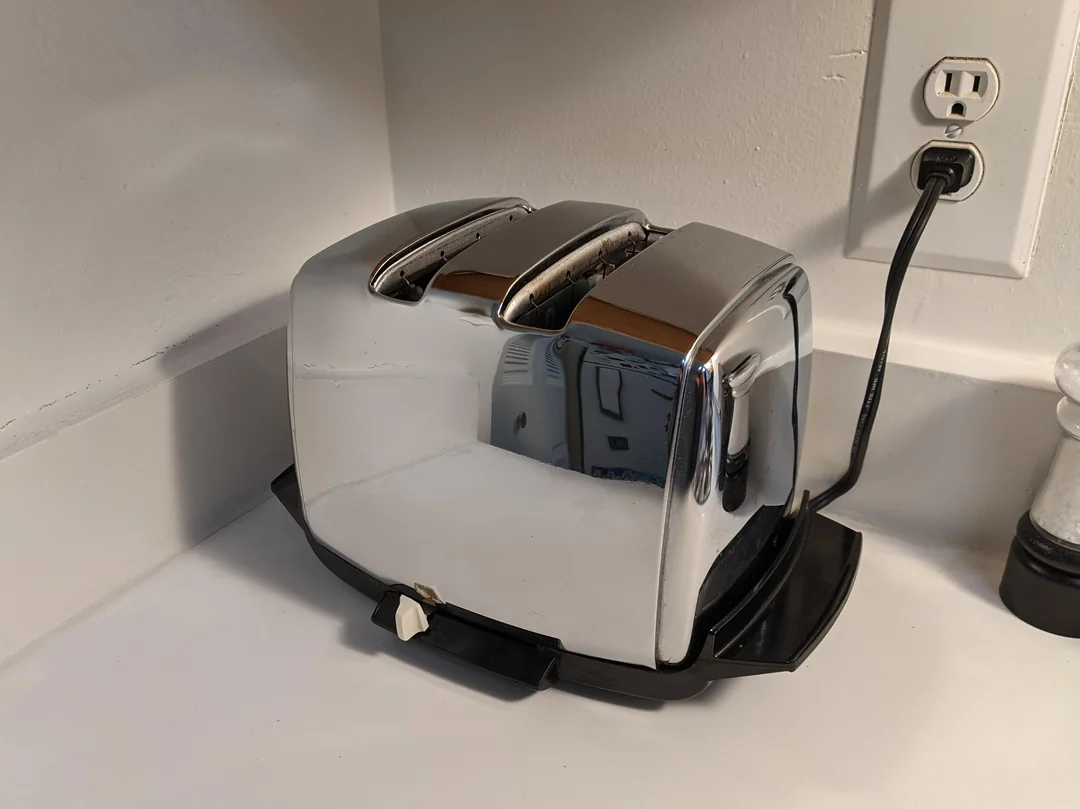 A shiny chrome toaster with two slots sits on a white kitchen counter near an electrical outlet. The toaster is plugged in, and a pepper grinder is visible to the right.