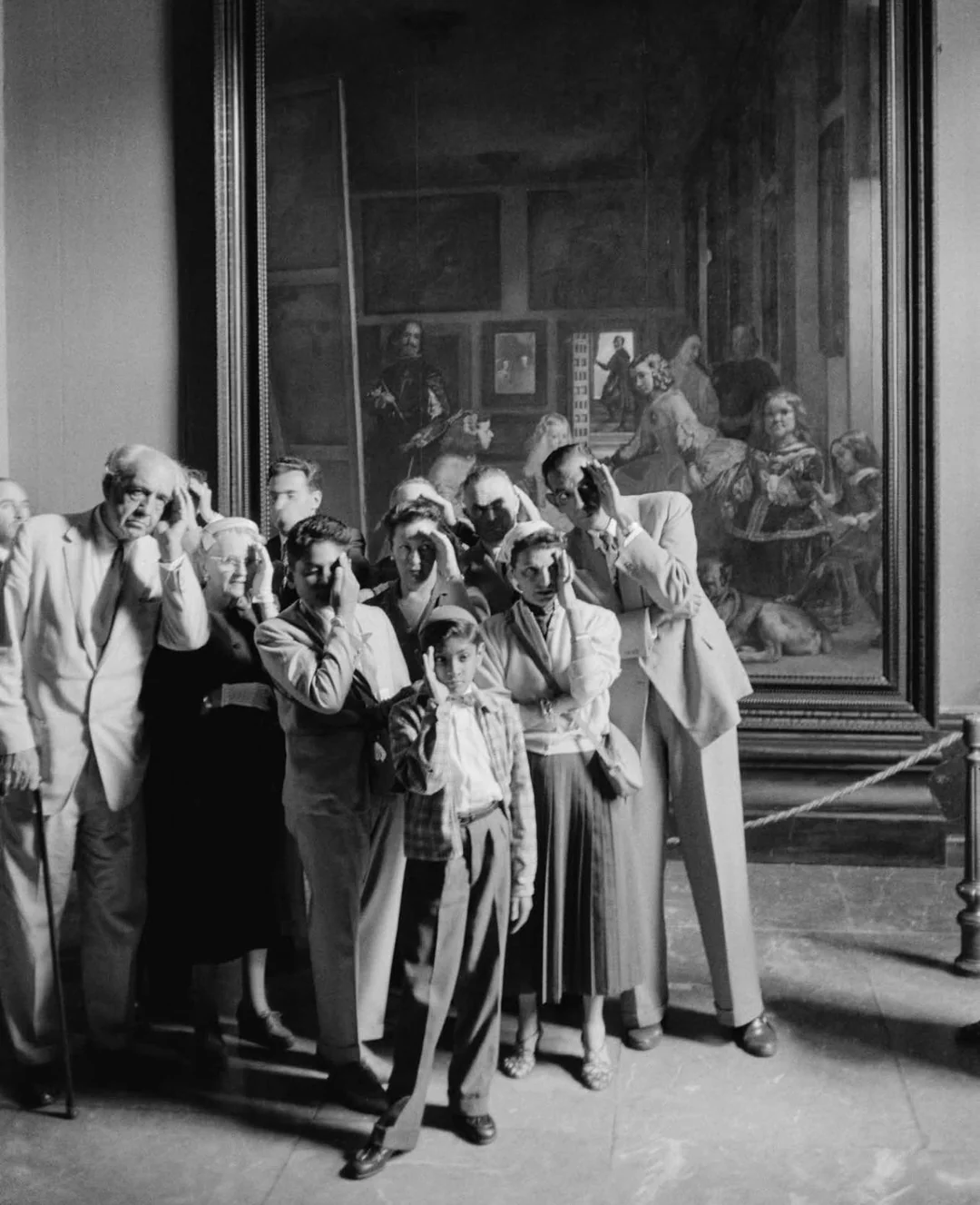 A group of people pose mimicking the gestures of figures in the large painting behind them at an art museum, creating a playful and lighthearted scene.
